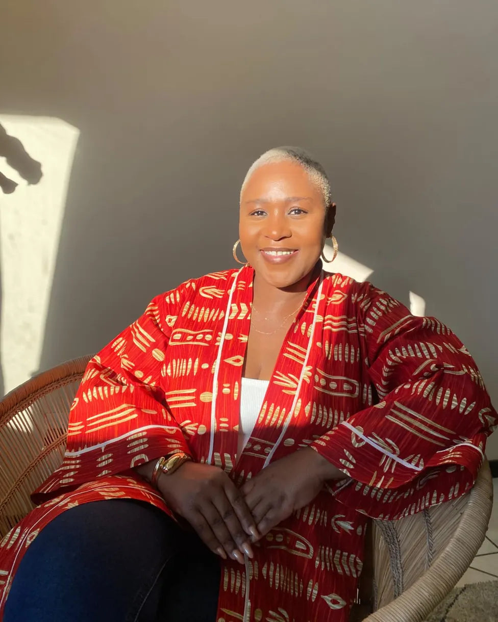 Cecilia Kapasula smiling, seated in a wicker chair in sunlight, wearing a red and gold printed jacket.
